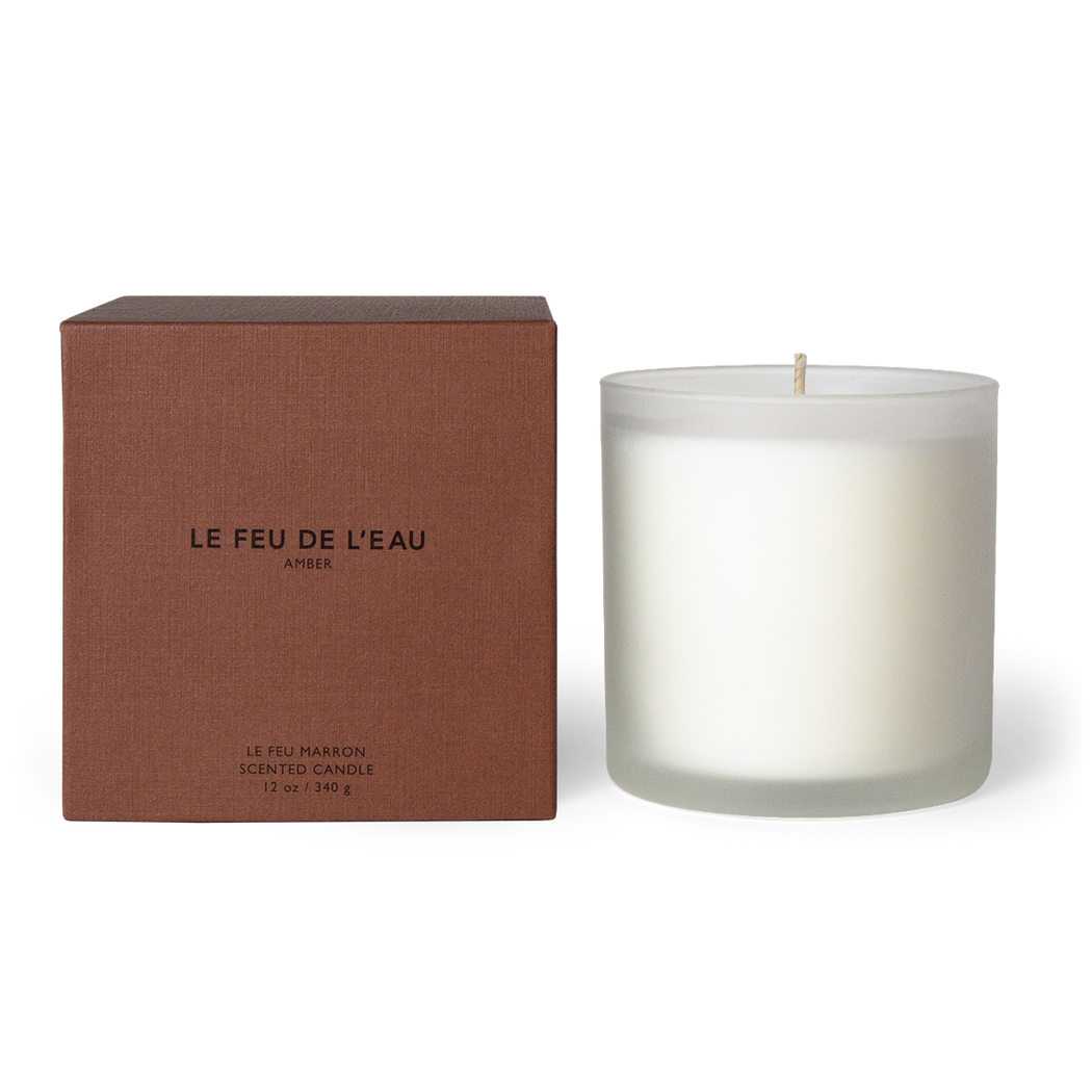 Indulge in the 12oz Marron candle, blending amber, sandalwood, patchouli, myrrh, resin, vanilla in Apricot and Coconut wax. With lead-free cotton wicks and a burn time of 65 hours, each candle brings a touch of artisanal luxury. Made in Los Angeles by LE FEU DE L'EAU and packaged in unique linen-textured paper. The candle is displayed unboxed, positioned beside its packaging, set against a clean white backdrop.