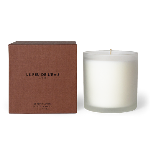 Indulge in the 12oz Marron candle, blending amber, sandalwood, patchouli, myrrh, resin, vanilla in Apricot and Coconut wax. With lead-free cotton wicks and a burn time of 65 hours, each candle brings a touch of artisanal luxury. Made in Los Angeles by LE FEU DE L'EAU and packaged in unique linen-textured paper. The candle is displayed unboxed, positioned beside its packaging, set against a clean white backdrop.