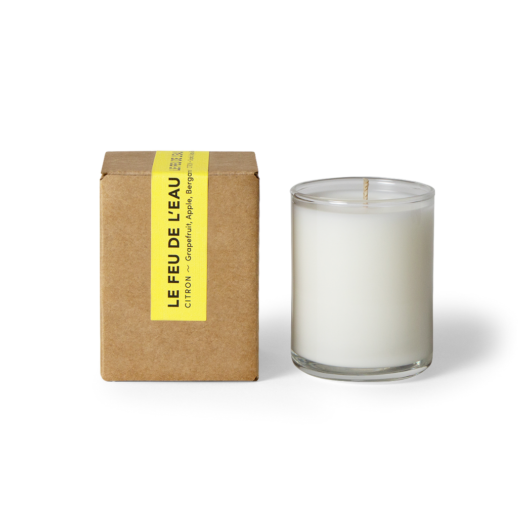 Discover the 3oz Citron glass candle, blending grapefruit, apple and bergamot- tangy grapefruit and clarifying apple welcome this new day. Hand-poured in LA, cruelty-free, with coconut apricot wax, natural fragrance, and lead-free cotton wicks. No harmful additives. The candle is displayed unboxed, positioned beside its packaging, set against a clean white backdrop.