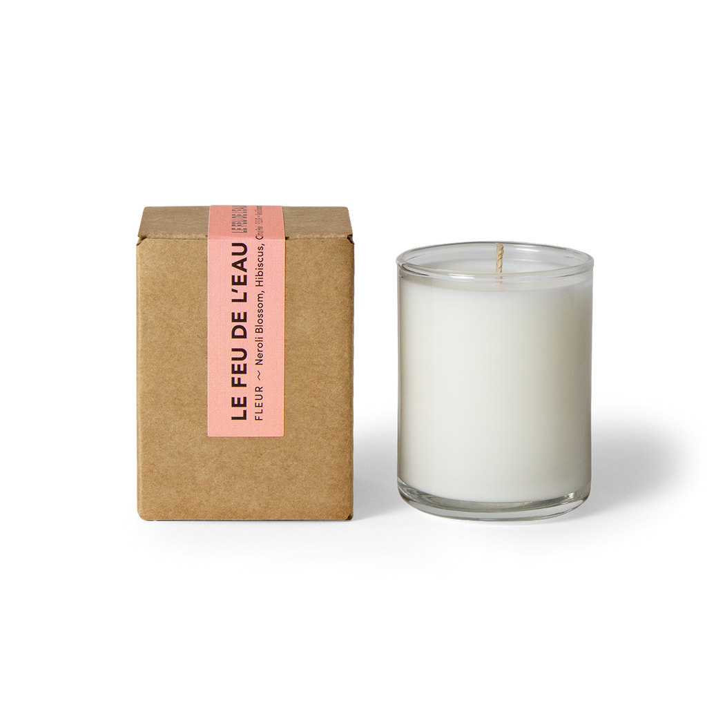 Discover the 3oz Fleur glass candle, blending neroli blossom, hibiscus, and citrus peel. Spring awakens with the liveliness of citrus cradled inside the warmth of neroli and ylang ylang. Hand-poured in LA, cruelty-free, with coconut apricot wax, natural fragrance, and lead-free cotton wicks. No harmful additives. The candle is displayed unboxed, positioned beside its packaging, set against a clean white backdrop.